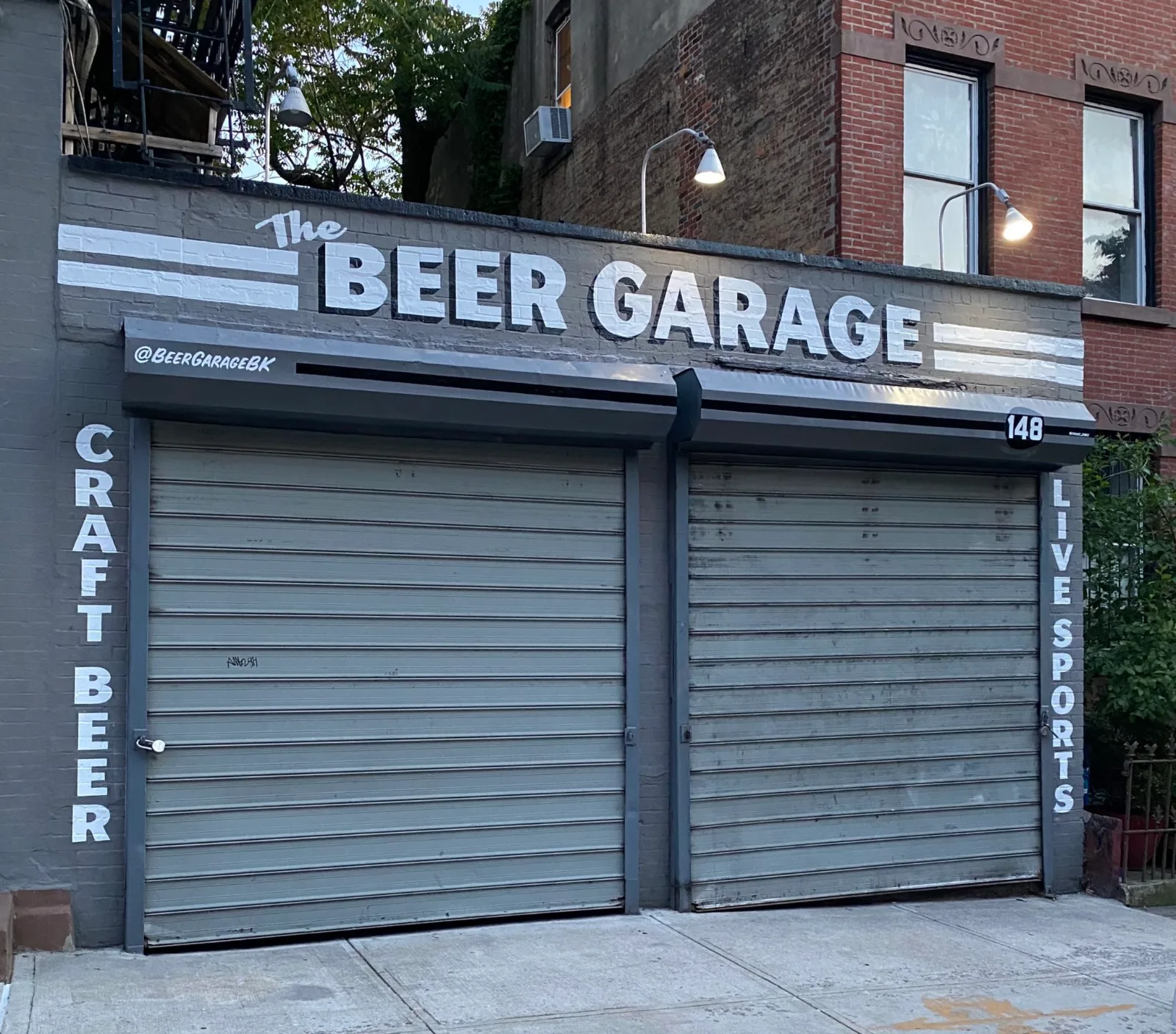 The corner of The Beer Garage at 148 5th Avenue in Park Slope, one of the bars near Barclays Center, showing the painted Beer Garage sign above the roll-down garage doors, the number 148 on the wall and the vertical CRAFT BEER and LIVE SPORTS signs on the Douglass Street side