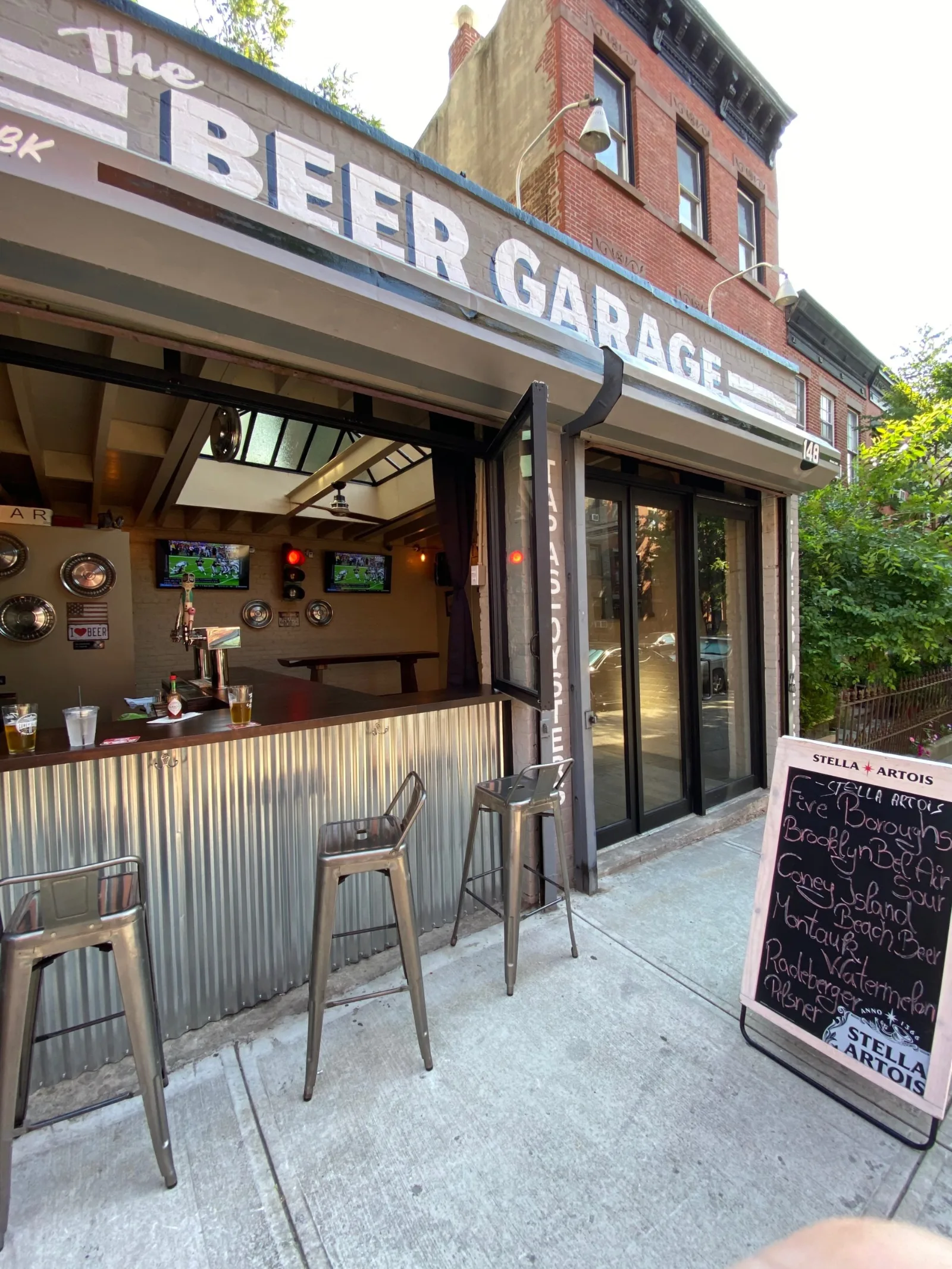 Metal stools at the open bar window of The Beer Garage, a cocktail bar in Park Slope option at 148 5th Avenue, with a chalkboard drinks board on the sidewalk