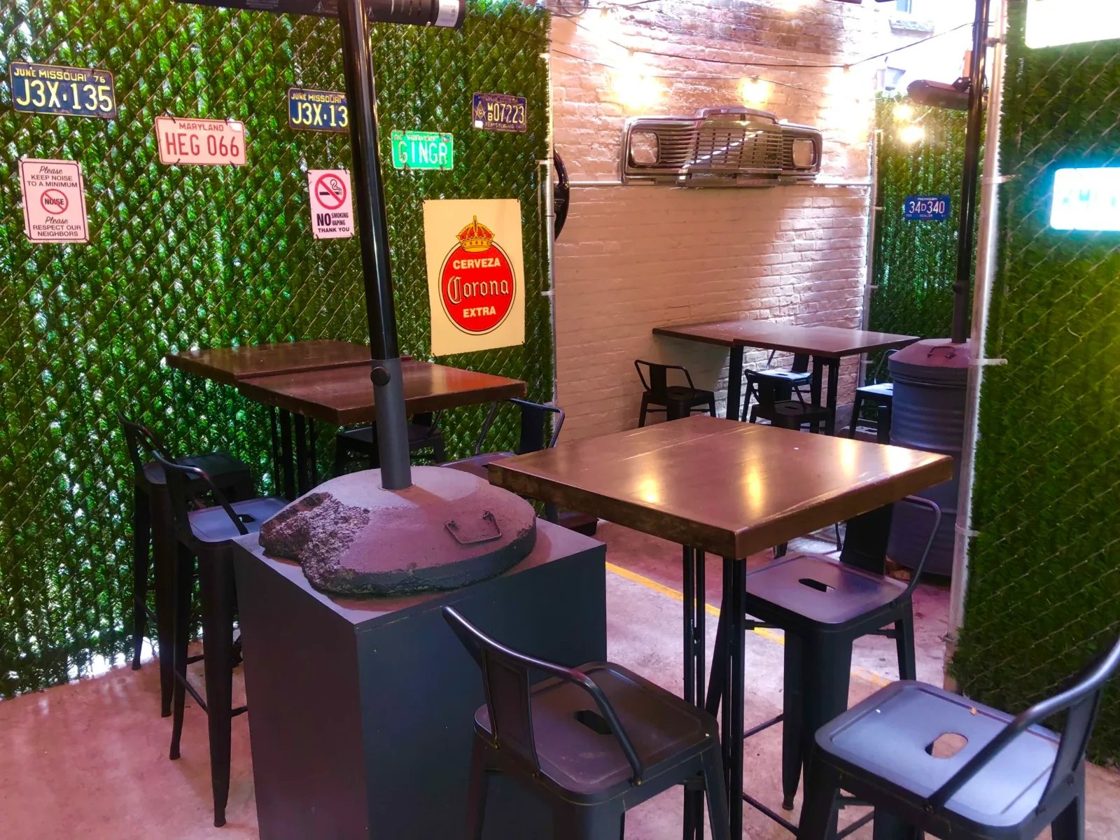 The heated backyard garden at The Beer Garage, a beer garden Manhattan locals can use all winter: license plates on green hedge walls, a heater pole, a car grille on white brick and high-top tables