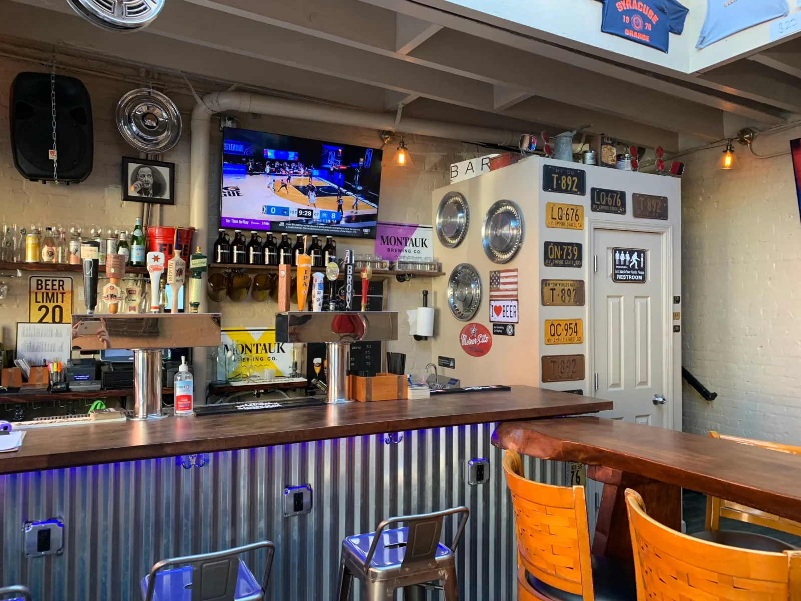 The bar at The Beer Garage in Park Slope, a holiday party venue in Brooklyn, with two draft towers, a television over the back bar, chrome hubcaps and old license plates on the wall, and a long wooden table with orange chairs