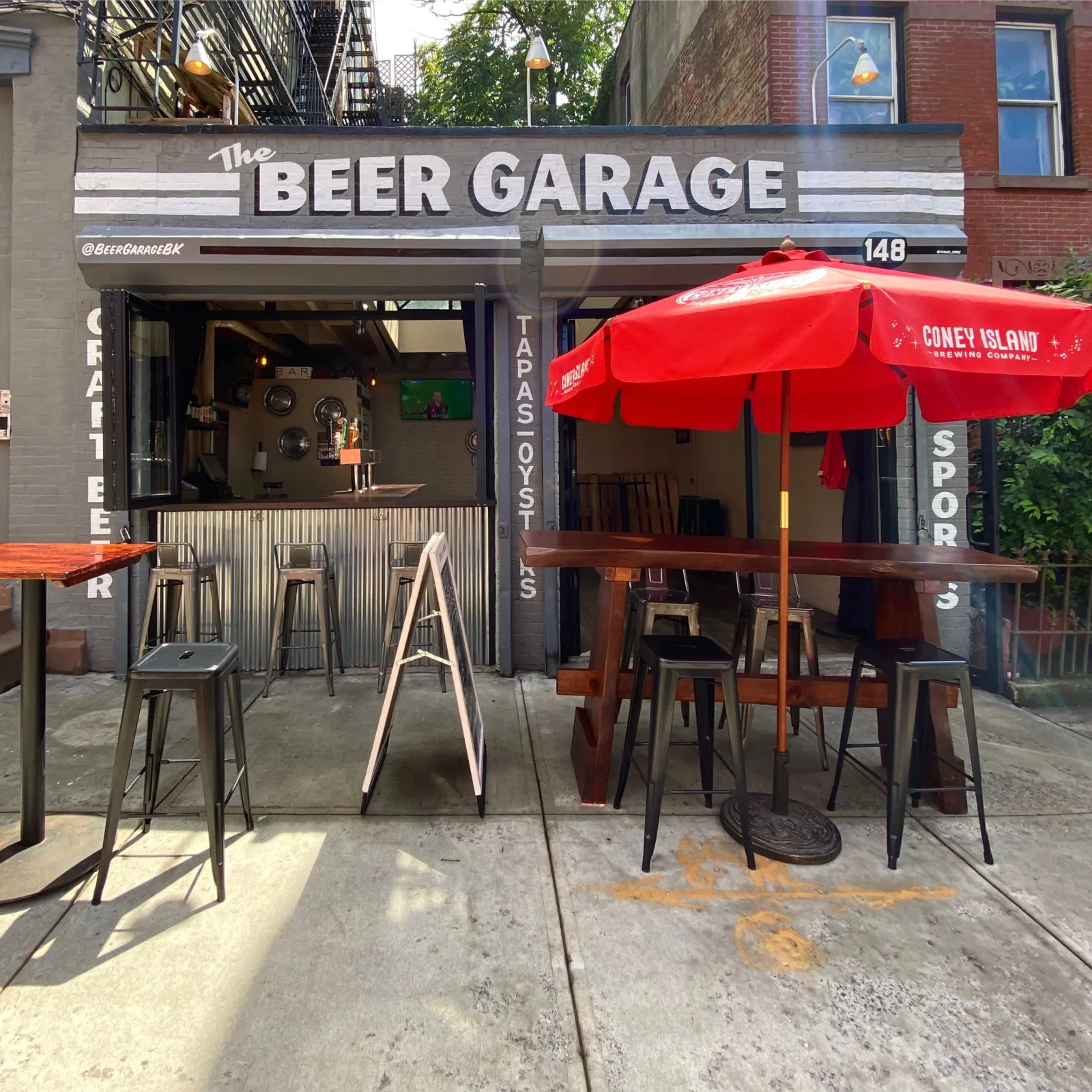 Sidewalk tables and stools under a red umbrella in front of The Beer Garage, a date night bar in Park Slope, with the garage door rolled up