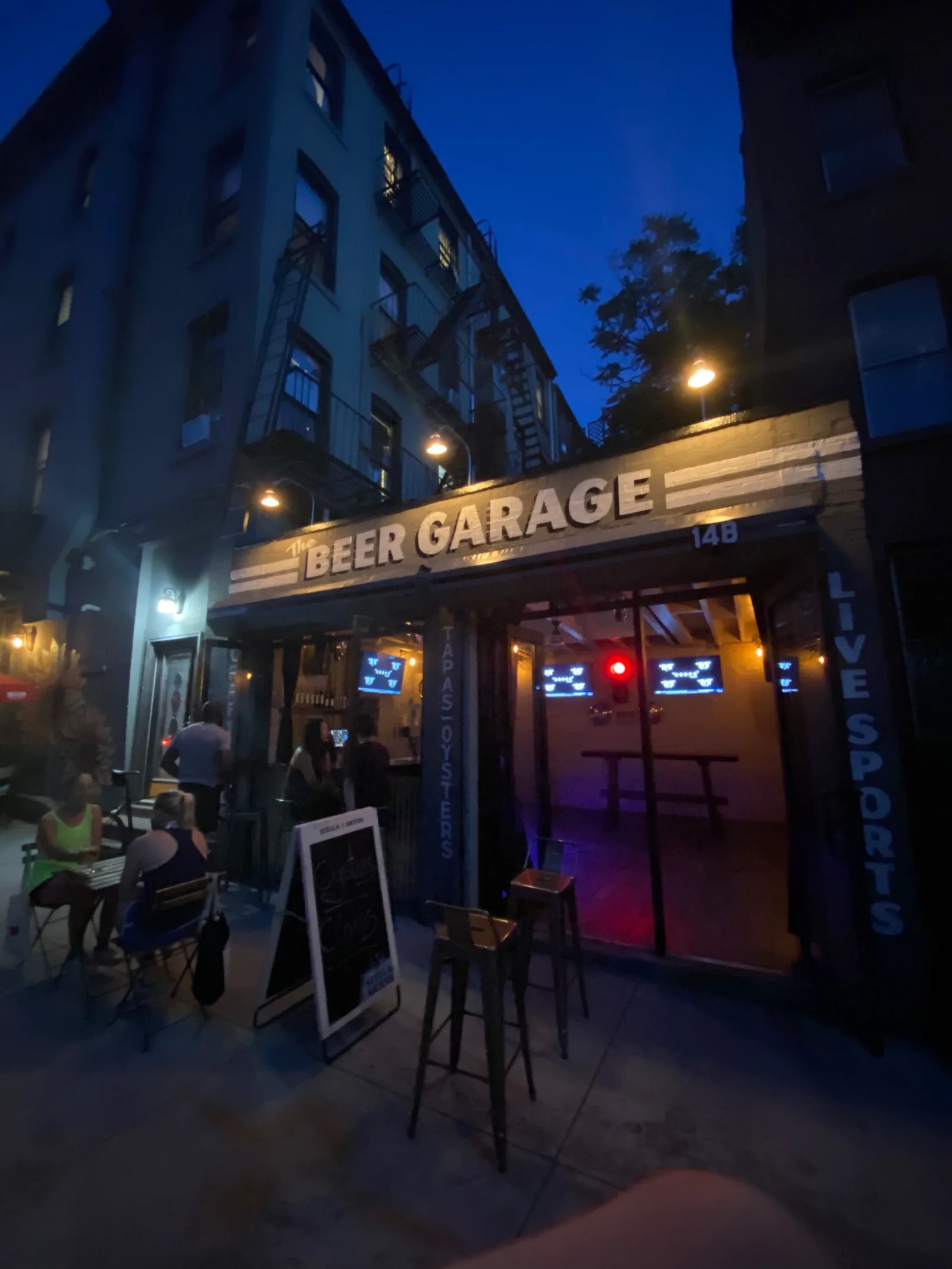 The Beer Garage at night at 148 5th Avenue, home of an oyster happy hour in Park Slope, with TAPAS and OYSTERS lettering on the pillar beside the open doors and LIVE SPORTS on the other side